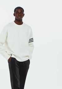 Sweatshirt blanc avec un design à trois rayures noires sur la manche. Il présente un col rond et un petit logo noir sur l'ourlet. Associé à un pantalon sombre.