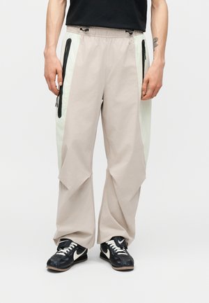 Άτομο που φοράει μπεζ παντελόνια με φαρδιά πόδια, λευκές πλευρικές ταινίες και μαύρες τσέπες με φερμουάρ, συνδυασμένα με μαύρα και λευκά παπούτσια Nike.