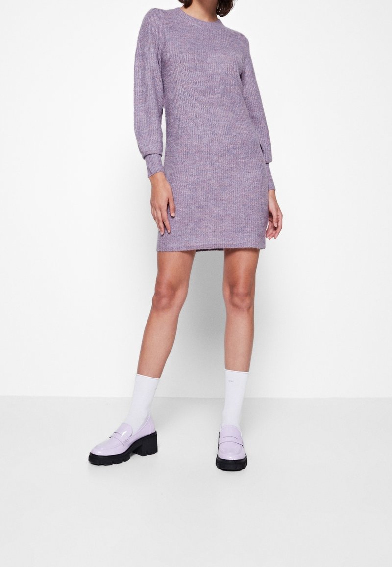 Femme portant une robe en maille lavande longueur genoux, manches longues, associée à des chaussettes blanches et des mocassins lavande épais, debout contre un mur blanc.