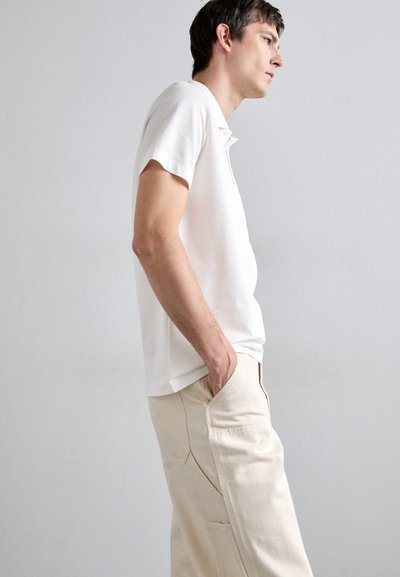White short-sleeve polo shirt with a classic collar, worn with light beige pants featuring patch pockets and a textured finish.