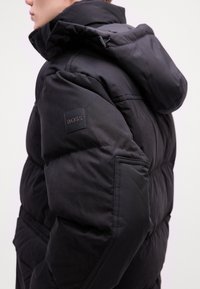 Black padded jacket with a hood, featuring a square BOSS logo on the sleeve. Smooth fabric and quilted texture enhance its warmth.