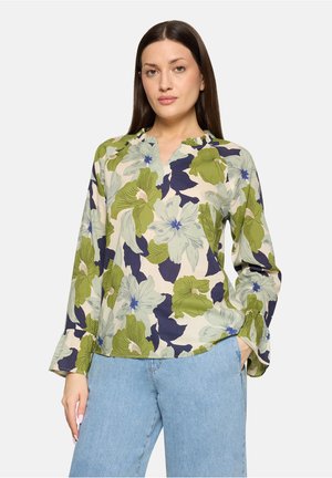 Femme aux longs cheveux foncés portant un chemisier à manches longues à motifs floraux verts et bleu marine, associé à un jean bleu clair.