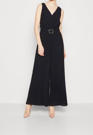 Mujer con mono negro plisado sin mangas, escote en V y cinturón en la cintura, combinado con zapatos de tacón altos en tono nude.