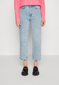 Hellblaue, hochgeschnittene Jeans mit geradem Bein, ausgestattet mit rohen Säumen und einem klassischen Fünf-Taschen-Design. Kombiniert mit schwarzen Mokassins.