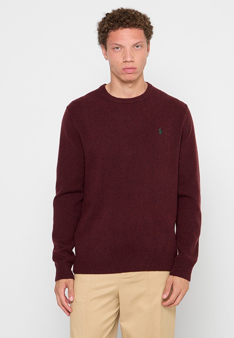 Pull en tricot bordeaux avec un col rond, des bords-côtes aux poignets et à l'ourlet, arborant un petit logo vert sur la poitrine gauche. Texture lisse, coupe régulière.