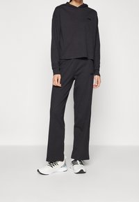 Black hoodie and matching sweatpants in soft fabric, featuring a relaxed fit. Accented with a logo on the chest and side of pants. White sneakers.