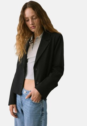 Blazer nero dal design aderente, con due bottoni e revers a collare, abbinato a un crop top grigio chiaro e jeans in denim azzurro chiaro.