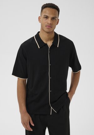 Jeune homme portant une chemise noire à manches courtes avec un bord beige, debout, une main dans la poche, sur fond uni.