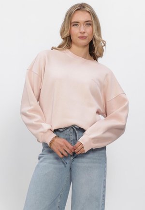 Ung kvinde med langt blondt hår iført en lyserød oversized sweatshirt og lyseblå højtaljede jeans, stående mod en hvid baggrund.