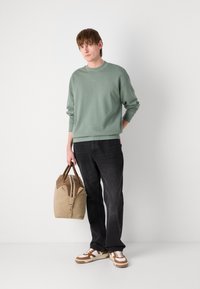 Sudadera verde claro, jeans negros y zapatillas marrones. Sosteniendo una bolsa de lona beige con detalles marrones y cremallera.