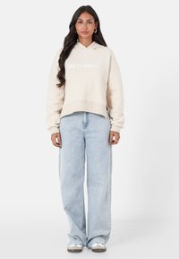 Kremowy, krótki hoodie z logo z przodu, w połączeniu z jasnoniebieskimi, szerokimi spodniami dżinsowymi i szarymi butami sportowymi. Gładki materiał i luźny krój.