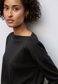 Sweatshirt noir avec un col rond, des accents côtelés aux épaules et une texture lisse. Visage du modèle partiellement visible avec un maquillage naturel.