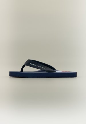 Chancla azul marino con tiras de tela negras que incluyen el logo blanco "Tommy Hilfiger" y un pequeño detalle de bandera roja, blanca y azul en la plantilla.