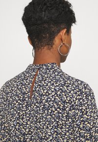 Blusa floreale blu navy con un piccolo dettaglio a apertura posteriore, caratterizzata da motivi di fiori bianchi e gialli su tessuto leggero. Collo rotondo e maniche corte.