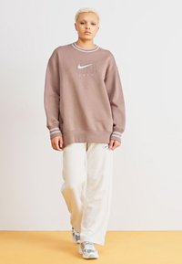 Barna Nike pulóver fehér logóval, bordás gallérral és mandzsettával; világosbézs, laza szabású nadrággal és szürke sportcipővel párosítva. Sima textúra.