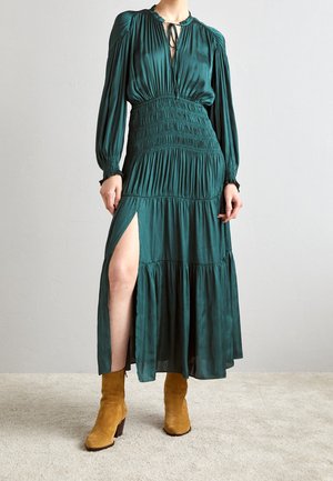 Kobieta w długiej sukience w kolorze morskim z marszczonym materiałem i rozcięciem do uda, w parze z musztardowymi zamszowymi botkami, stojąca na dywanie.