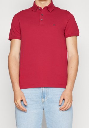 Mann trägt ein rotes Polohemd mit kurzen Ärmeln und Knöpfen, kombiniert mit hellblauen Jeans, steht vor einem schlichten Hintergrund.