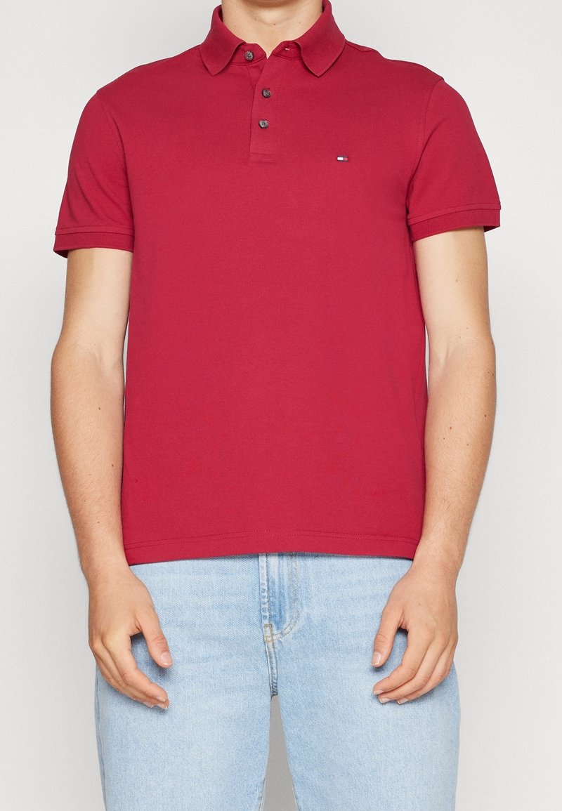 Homme portant un polo rouge à manches courtes avec des boutons, associé à un jean denim bleu clair, debout devant un fond uni.