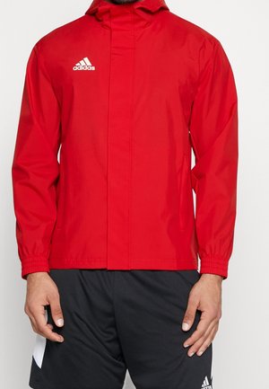 Hombre con chaqueta roja de Adidas con capucha y pantalones cortos negros de Adidas, de pie con las manos relajadas a los costados.