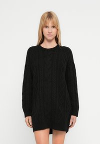 Zwarte gebreide trui jurk met kabelgebreide structuur, lange mouwen en een geribde halslijn en zoom. Casual pasvorm met zijsplitten.