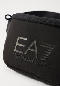 Černá nylonová taška přes pás s embosovaným logem EA7, texturovanou tkaninou a zipovým uzávěrem. Kompaktní a aerodynamický design pro snadné nošení.