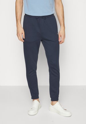 Pantalons de jogging navy avec une taille élastique et un cordon de serrage. Dotés de poignets côtelés et d'un logo discret. Associés à des baskets blanches.