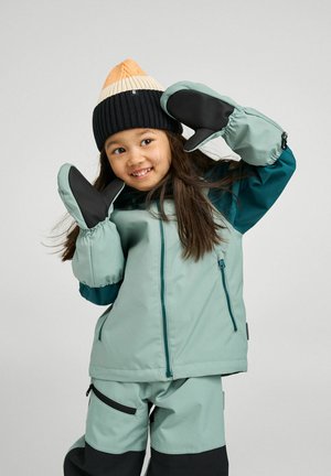 Małe dziecko uśmiechające się, ubrane w turkusową zimową kurtkę i spodnie, czapkę z dzianiny w czarnym i pomarańczowym kolorze oraz za duże turkusowe rękawiczki, pozujące z uniesionymi rękami.