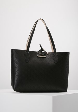 Sac cabas noir Guess avec motif de logo embossé, deux longues anses et une petite étiquette métallique décorative sur un socle blanc.