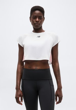 Donna con capelli corti che indossa un top sportivo bianco corto e leggings neri a vita alta, in piedi contro uno sfondo chiaro semplice.