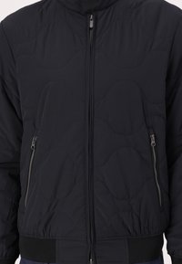 Zwarte gewatteerde jas met hoge kraag, voorrits en twee zijzakken. Voorzien van elastische manchetten en onderzoom voor extra warmte.