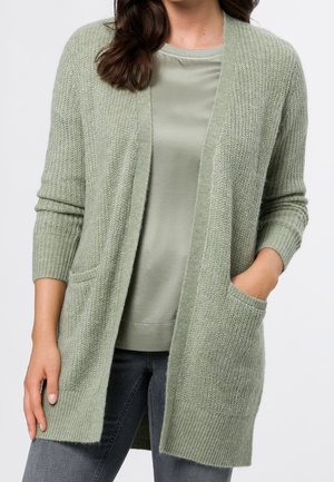 Femme portant un cardigan en maille vert clair avec poches sur une chemise verte assortie et un jean foncé, main dans la poche, sur fond uni.