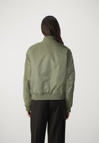 Zelená bomber bunda s hladkou, lesklou textúrou; rebrovaný golier a manžety; priliehavý pás; obsahuje bočné vrecká a prešité akcenty.