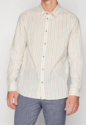 Mężczyzna noszący beżową koszulę z długim rękawem z cienkimi pionowymi paskami i brązowymi guzikami, w połączeniu z szarymi teksturowanymi spodniami.