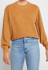 Kvinna som bär en senapsgul ribbad stickad tröja med ballongärmar, ihop med högmidjade ljusblå jeans mot en vit bakgrund.