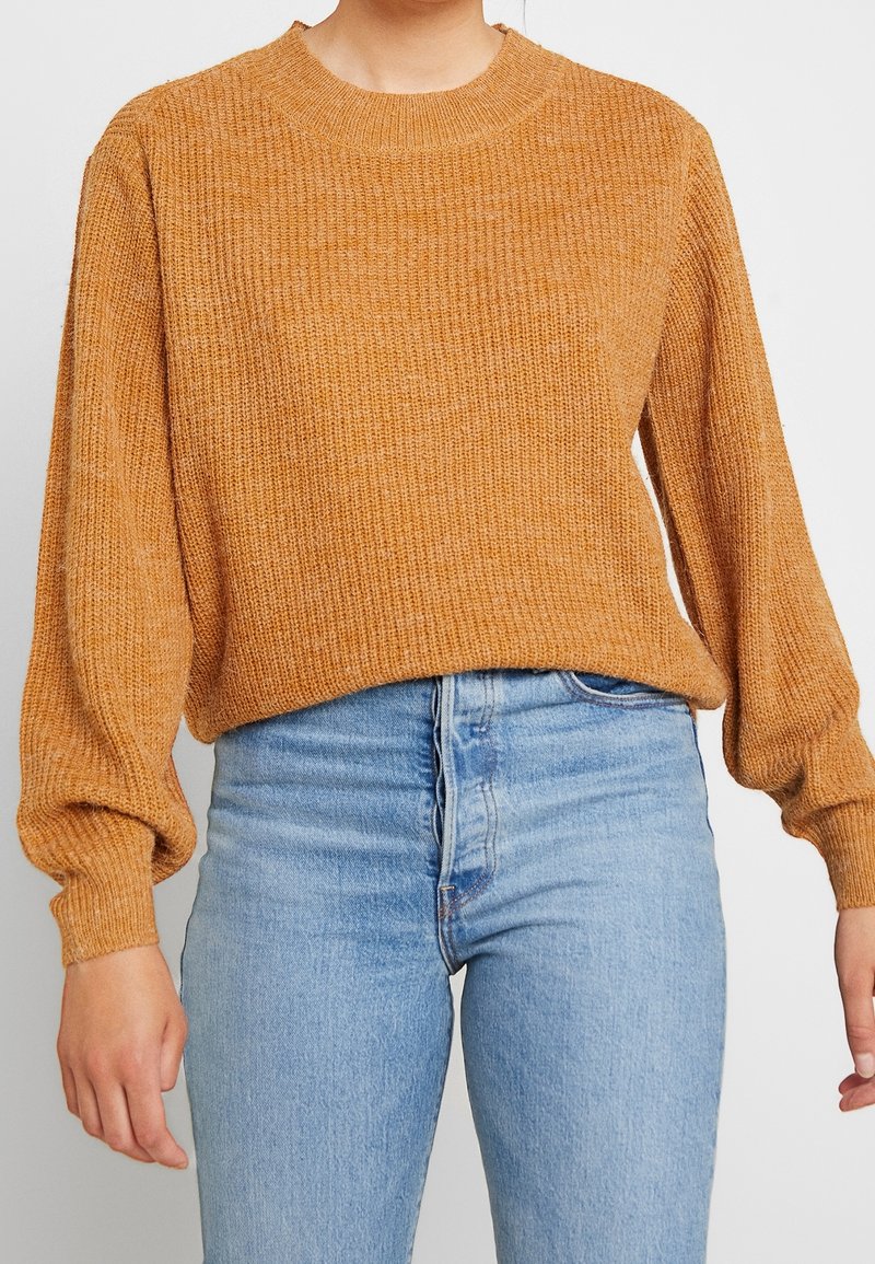 Kvinna som bär en senapsgul ribbad stickad tröja med ballongärmar, ihop med högmidjade ljusblå jeans mot en vit bakgrund.