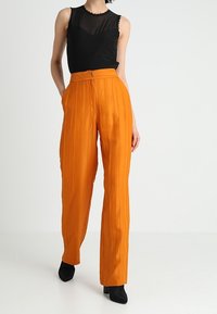 Femme portant un pantalon plissé orange taille haute, un haut sans manches noir avec un col en tissu transparent, et des bottines noires, se tenant contre un fond uni.