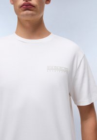 Bílý bavlněný tričko s malým embosovaným logem na levé části hrudi. Design zahrnuje klasický kulatý výstřih a krátké rukávy.