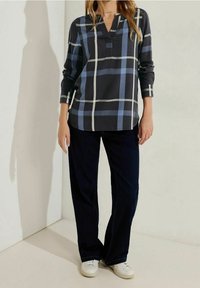 Femme portant une chemise noire à manches longues avec un motif à carreaux bleu et blanc, un pantalon large et sombre, et des baskets blanches, debout contre un mur clair.