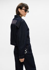 Σκούρο τζάκετ από τζιν με κεντημένη την λέξη "HEROES" στην πλάτη, συνδυασμένο με σκούρες τζιν παντελόνες που διαθέτουν διακοσμητική ραφή και λογότυπο.