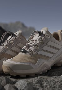 Beżowe buty trekkingowe o teksturowanej powierzchni, z systemem blokady sznurowadeł, wzmocnionym czubkiem i białymi akcentami po bokach. Wytrzymała podeszwa zapewniająca przyczepność.