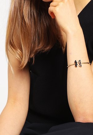 Femme portant un haut noir sans manches avec un bracelet en or orné de breloques en forme de moustache noire et de lunettes à son poignet.