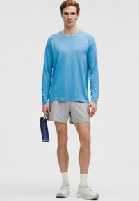 Vaaleansininen pitkähihainen urheilupaita teksturoidulla kankaalla, harmaat shortsit, valkoiset sukat ja vaaleat kengät. Pitelee sinistä vesipulloa.