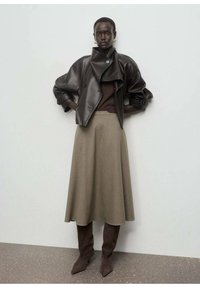 Veste en cuir marron courte, pull marron foncé, jupe mi-longue verte olive et bottes pointues marron. Textures lisses avec un look superposé.