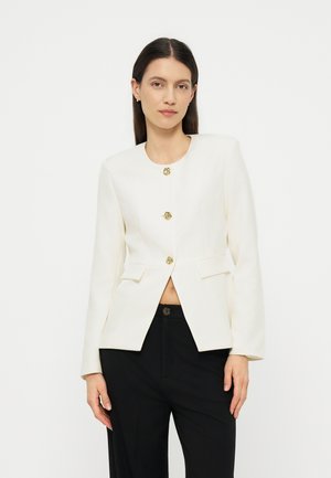 Kvinde med glat mørkt hår iført en hvid blazer med guldknapper og sorte bukser, stående mod en hvid baggrund.