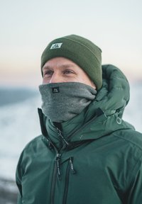 Giubbotto puffer verde con colletto alto, scaldacollo grigio e berretto lavorato a maglia verde scuro. Presenta dettagli con cerniera e un'etichetta con logo.