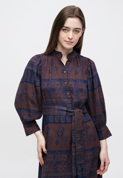 Νέα γυναίκα με μακριά καστανά μ�αλλιά, ντυμένη με φόρεμα καφέ και ναυτικό μπλε με λουλουδάτο μοτίβο, φουσκωτά μανίκια και ζώνη στη μέση.