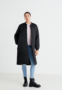 Crni quilted dugi kaput sa stajaćim ovratnikom, nošen preko svijetloružičastog pulovera, uparen sa plavim trapericama i crnim čizmama na vezivanje.