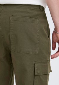 Oliivinvihreät cargo-housut, joissa on sileä puuvillainen pinta ja jotka sisältävät takataskun sekä sivulla olevan cargo-taskun säilytystarpeita varten. Yksinkertainen, käytännöllinen muotoilu.