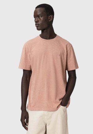 Mężczyzna ubrany w wyblakły różowy T-shirt z krótkim rękawem i jasnobeżowe spodnie, patrzący w lewo, z jedną ręką w kieszeni.