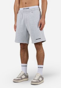 Calções de algodão cinzentos com cintura elástica, bolsos laterais e detalhe em preto do logótipo na coxa esquerda, combinados com ténis claros e meias brancas.